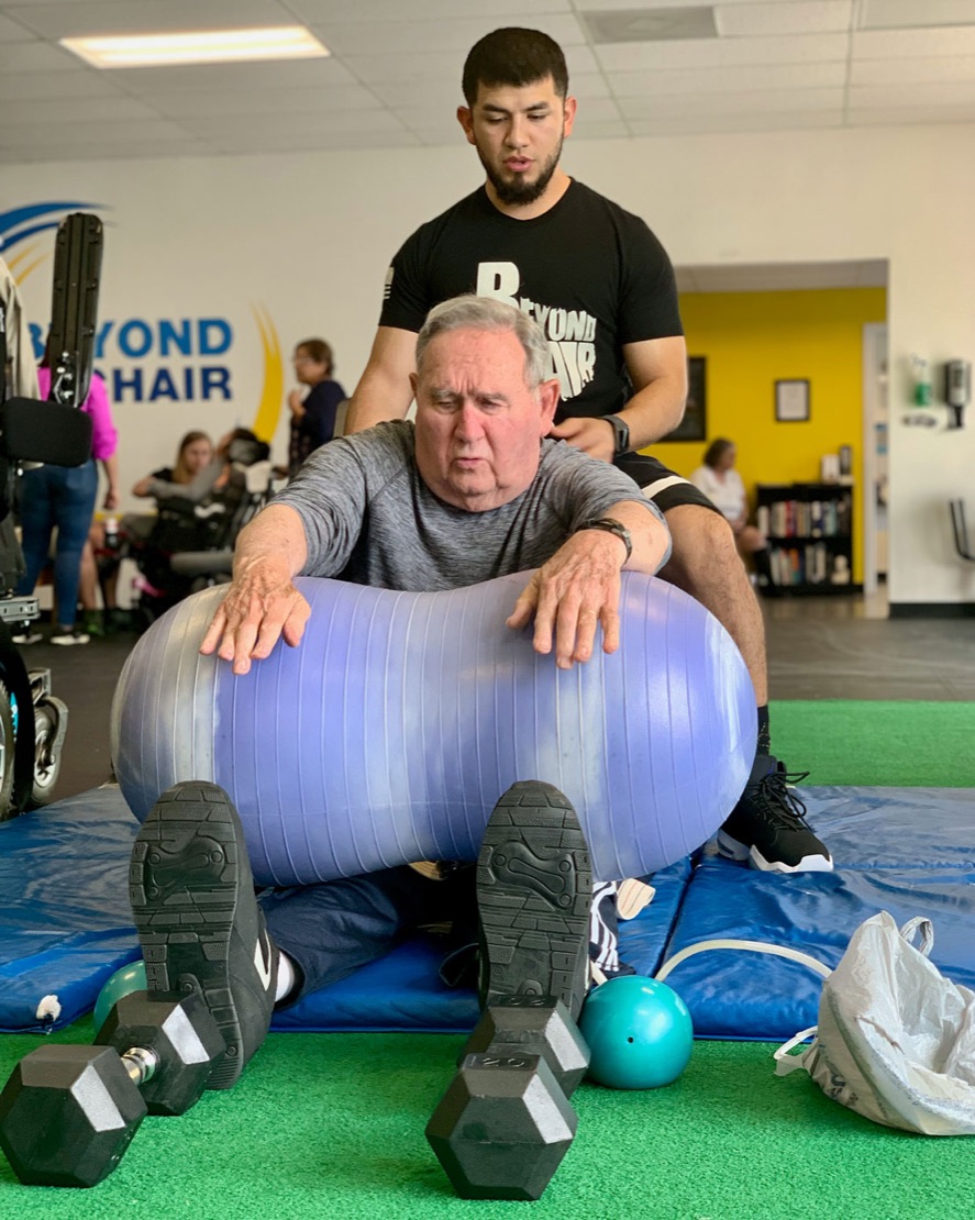 A trainer stands behind an older member who sits on a mat and balances over a large exercise ball.