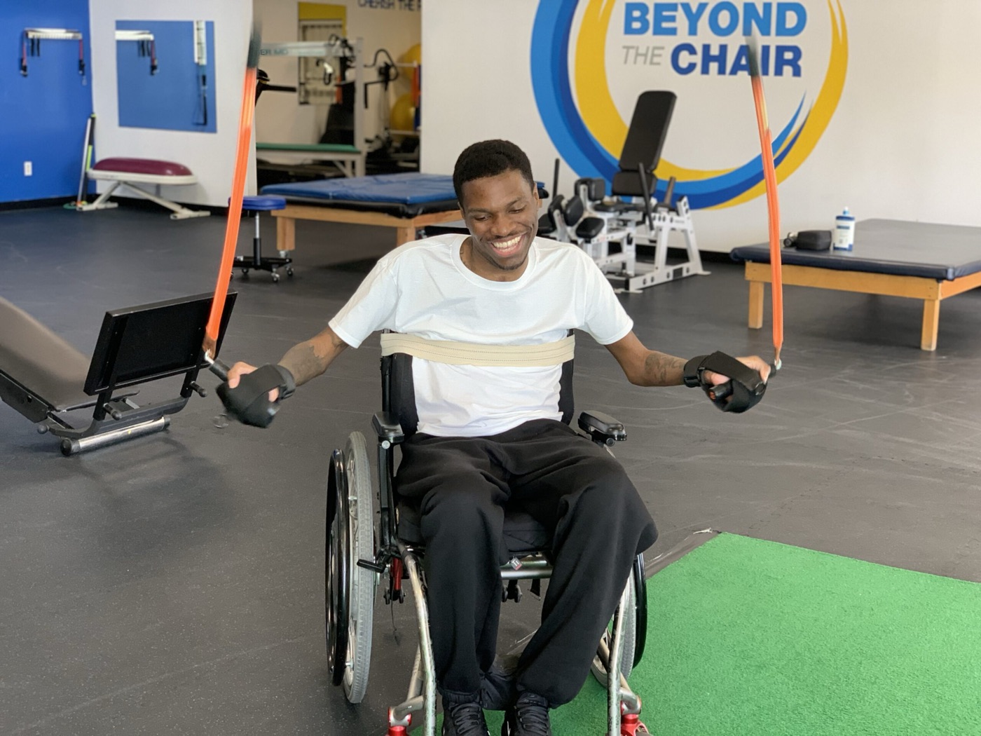 A smiling member in a wheelchair pulls resistance bands in front of the logo wall.