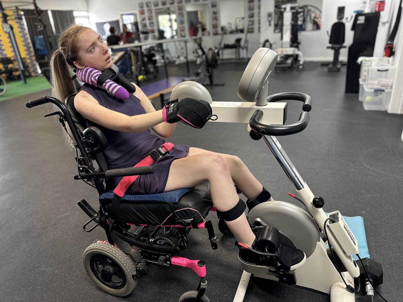 A young woman in a pink wheelchair pedals an arm-and-leg cycle.