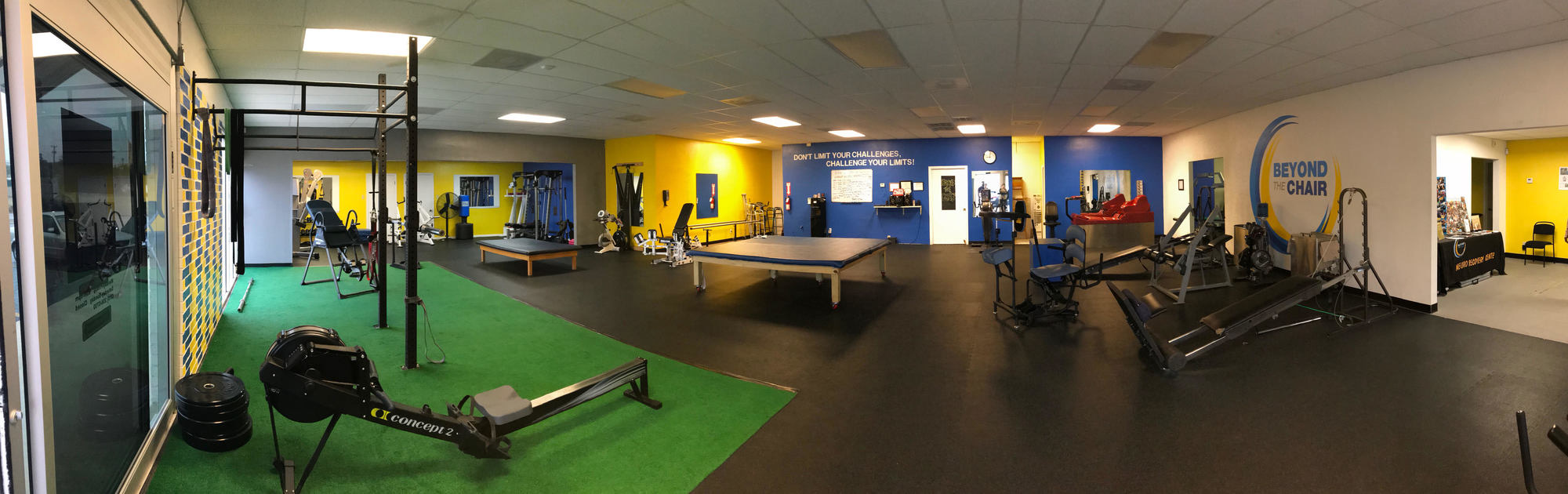The Beyond The Chair gym floor: turf, a rowing machine, treatment tables, and standing and gait equipment in front of yellow and blue walls.