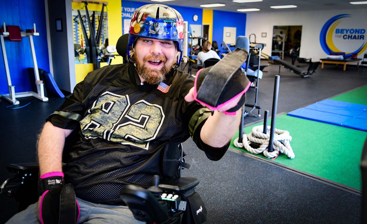 A grinning member in a decorated helmet and football jersey reaches a gloved fist toward the camera in the gym.