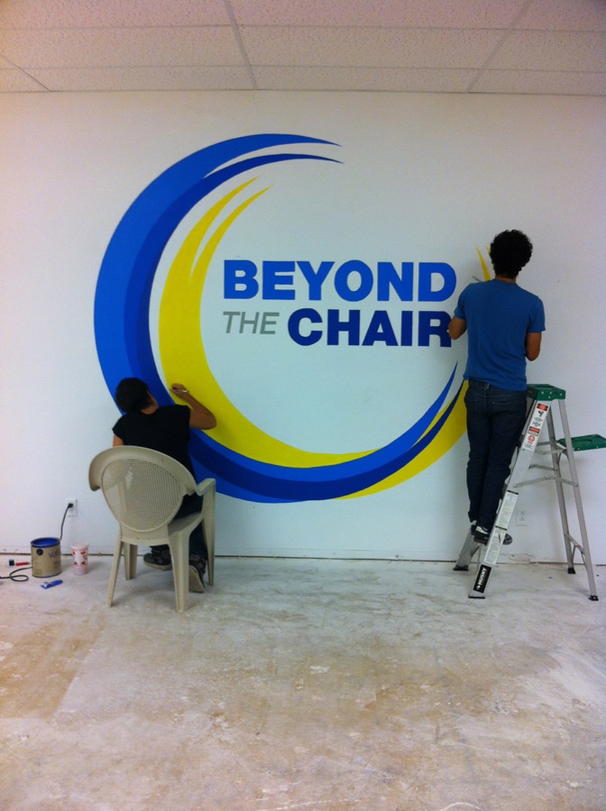 Volunteers painting the yellow and blue Beyond The Chair logo on a white wall.