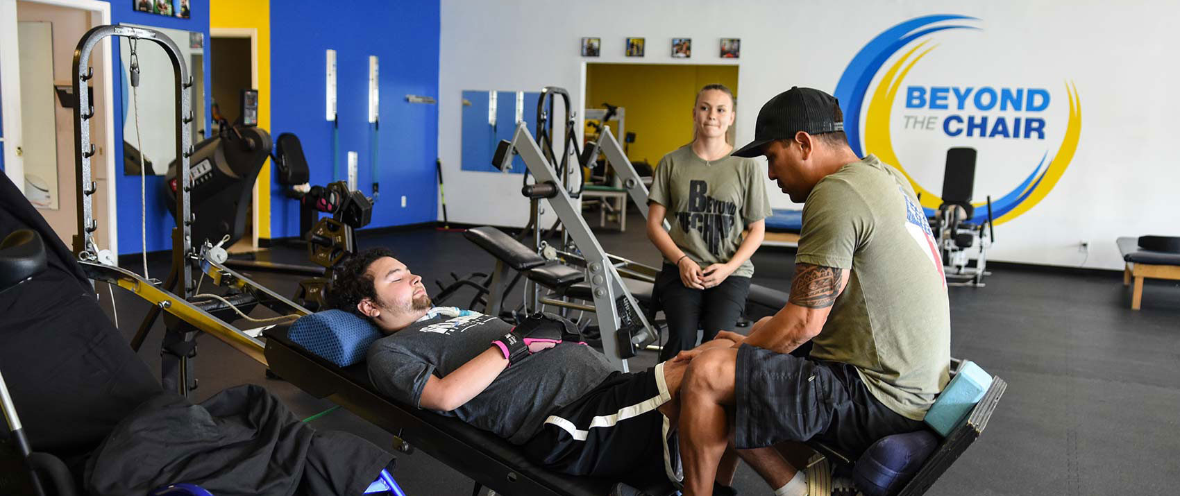 A member lies on a leg-press machine while a trainer works with his legs and another trainer watches.