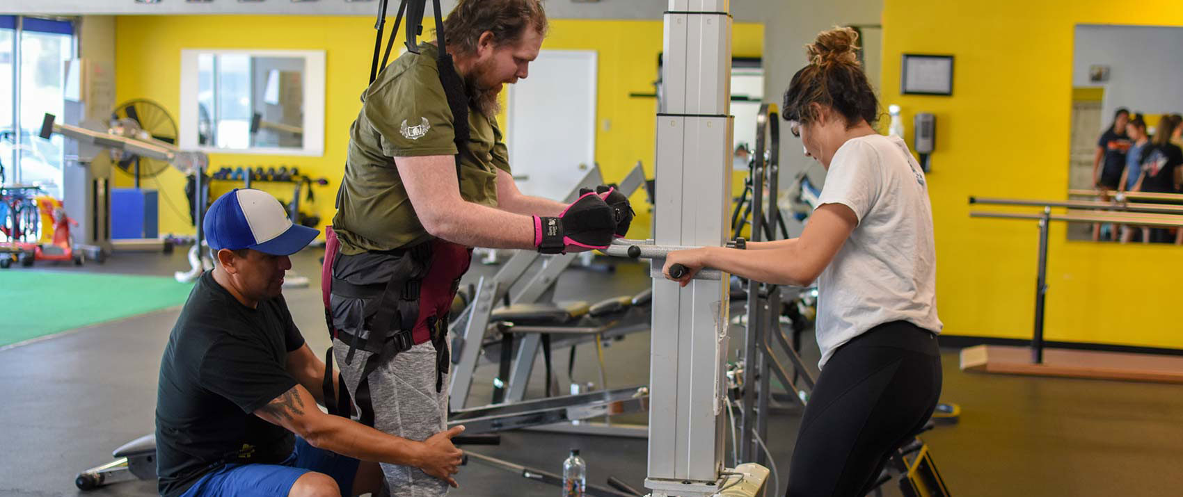 A member stands in a support harness at the gait station while two trainers guide his legs and the frame.