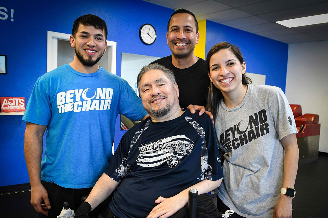 A smiling member in a wheelchair with three Beyond The Chair staff standing behind him in front of a blue wall.